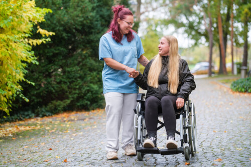 Schülerin schiebt Patientin im Rollstuhl und unterhält sich mit ihr